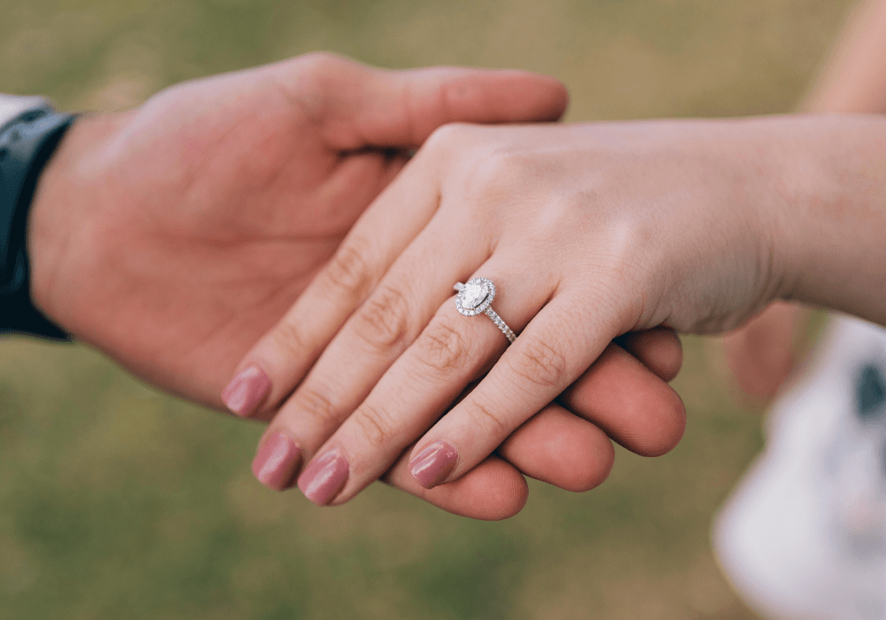 Hand wearing a diamond engagement ring with halo setting held outdoors